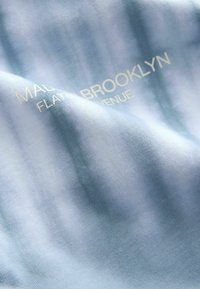 Világoskék szövet, amelyen halvány fehér betűkkel részben olvasható a "MAD BROOKLYN FLATS AVENUE" felirat.