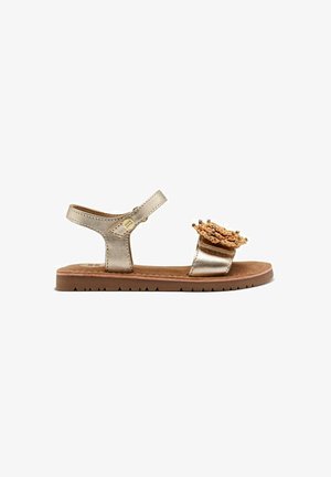 Metallische gouden sandaal met een gestructureerde bovenkant en decoratieve bloemaaccent. Voorzien van een verstelbare band en een platte rubberen zool.