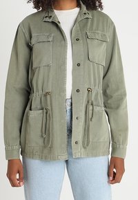 Olive green utility jacket with two chest pockets, drawstring waist, and button closures. Fabric appears textured and durable.