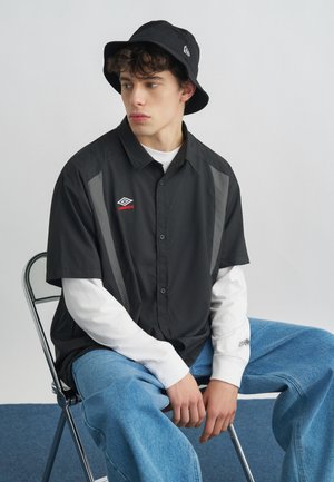 Mladi moški z rjuhasto črno klobučasto kapo, črno kratkimi rokavi Umbro majice preko belih dolgih rokavov in širokimi modrimi kavbojkami sedi na kovinskem stolu.