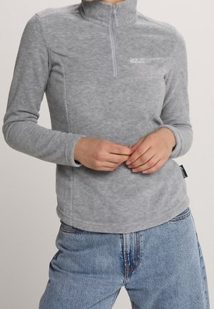 Personne portant un haut en polaire gris à manches longues avec une fermeture éclair partielle et un jean bleu clair, les mains jointes à la taille.