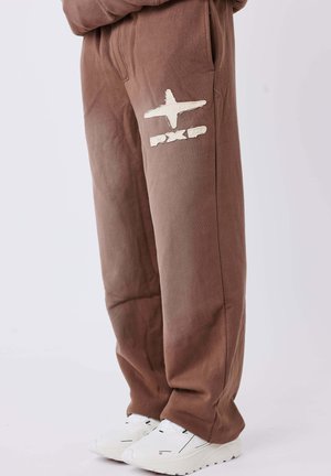 Pantalones de chándal marrones de algodón con bolsillos laterales, que presentan un logo blanco y detalles gráficos. Combinados con zapatillas blancas para crear contraste.
