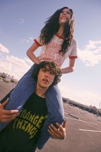 Δύο άτομα σε εξωτερικό χώρο, το ένα καθισμένο στους ώμους του άλλου. Το άτομο από κάτω φοράει μαύρο μπλουζάκι με κίτρινο κείμενο «STRANGE THINGS». Το άτομο πάνω φοράει άσπρο πουκάμισο με κόκκινη επένδυση και το κείμενο «WELCOME TO THE UPSIDE DOWN» — έχει μακριά σκούρα μαλλιά και γυαλιά ηλίου. Και οι δύο βρίσκονται σε χώρο στάθμευσης με έναν εν μέρει συννεφιασμένο ουρανό.