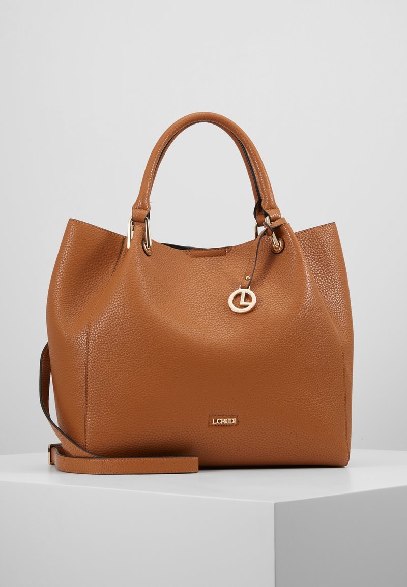 Sac à main en cuir texturé marron avec double poignée, une bandoulière amovible et un petit charme rond en or suspendu près du haut.