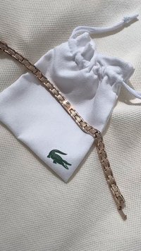 Rožinio aukso apdaila metalinis apyrankė, su sujungtais segmentais, šalia balto maišelio su žaliu krokodilo logotipu. Tekstūruota fono paviršius.