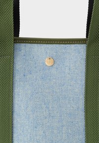 Světlomodrý textilní panel s jedním zlatým patentem, lemovaný zelenými texturovanými popruhy s viditelným stehem.