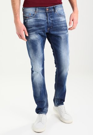 Homme portant un jean slim bleu délavé avec une légère usure et des baskets blanches, debout devant un fond clair uni.