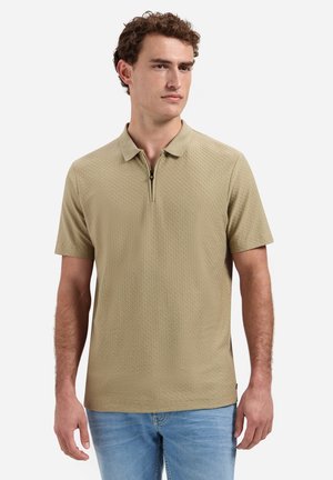 Mann trägt ein beiges kurzärmeliges, strukturiertes Poloshirt mit Frontreißverschluss, kombiniert mit hellblauen Jeans, stehend vor weißem Hintergrund.