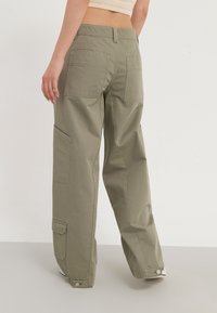Kaki kalhoty olivově zelené barvy s volnějším střihem, s několika kapsami, rovnými nohavicemi a přezkami na kotnících.