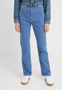 Personne portant un pantalon en velours côtelé bleu, des baskets blanches avec des semelles en gomme, et une veste en denim courte, debout sur un fond blanc uni.