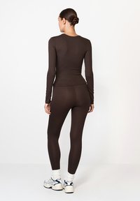 Barna hosszú ujjú felső és leggings, testhezálló fazon, rugalmas anyag, sima textúra. A modell sportcipőt visel, haja hátra van kötve.