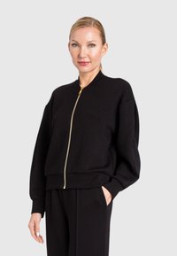 Felpa nera con zip, colletto alto, vestibilità comoda e polsini a costine. Presenta dettagli in oro e tasche laterali. Design semplice.