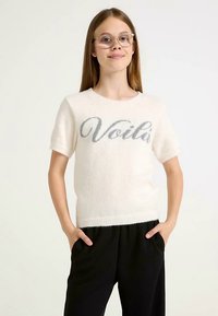 Maglione bianco a maniche corte realizzato in materiale morbido e soffice, con la parola "Voilà" in corsivo grigio sulla parte frontale.