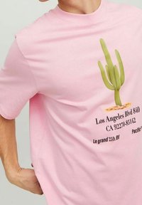 T-shirt rose à manches courtes présentant un graphique de cactus avec des détails textuels en dessous. Le matériau semble doux et a une texture semblable à celle du coton.
