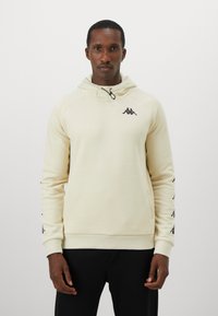 Man wearing off-white hoodie with black logo on chest and sleeves, standing against plain light background.