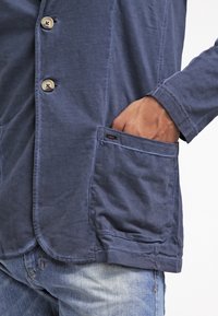 Uomo con giacca blu abbottonata e mano infilata nella tasca anteriore, abbinata a jeans di denim azzurro.