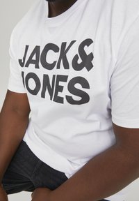 Bijela pamučna majica sa podebljanim crnim natpisom "JACK & JONES." Okrugli izrez, kratki rukavi i opušteni kroj. Nosite uz tamne traperice.