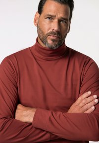 Maglione a collo alto a maniche lunghe di colore ruggine con una texture liscia, caratterizzato da un design aderente e uno stile sobrio e minimalista.