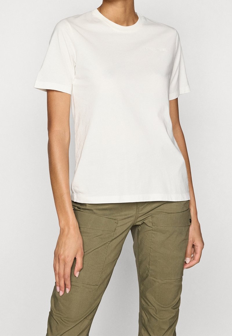 Femme portant un t-shirt blanc uni à manches courtes et un pantalon cargo vert olive, debout devant un fond uni.