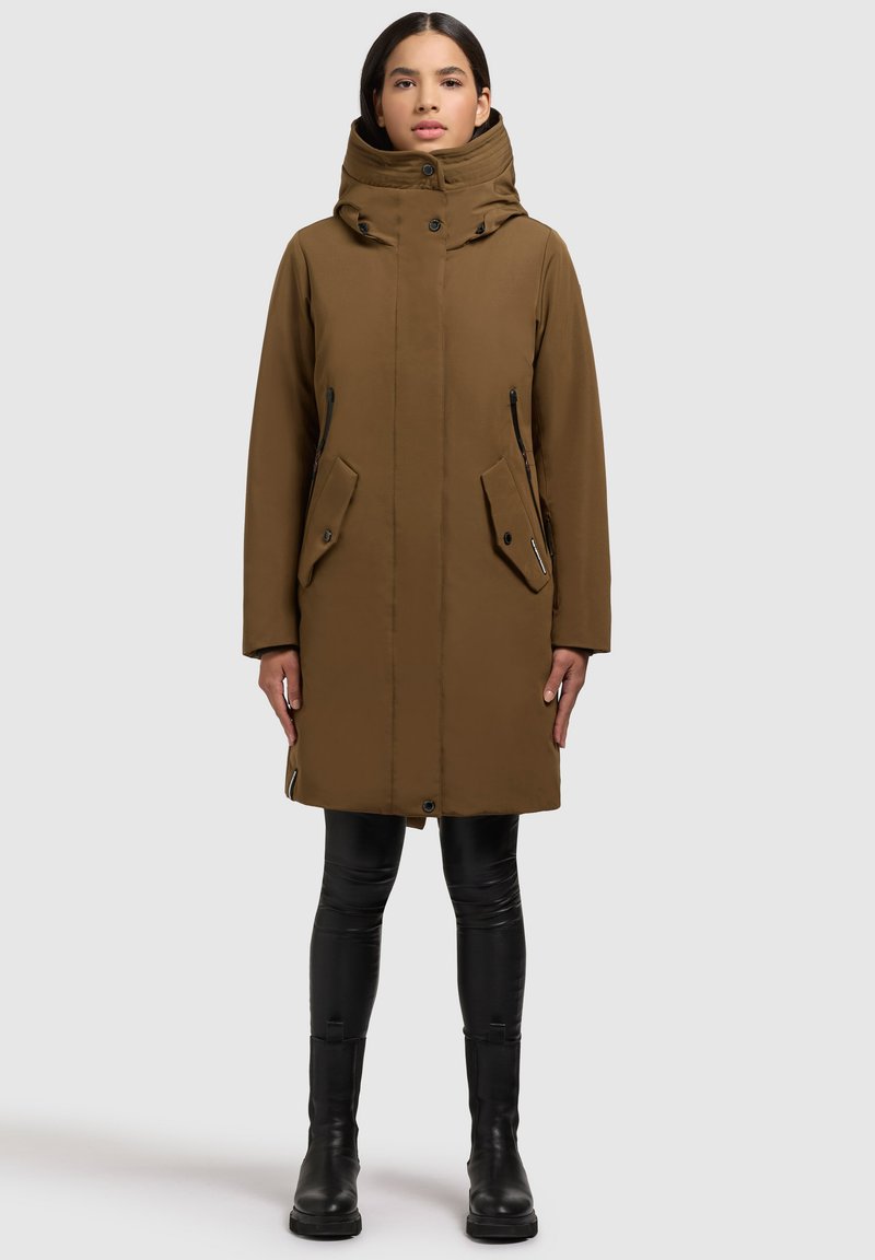 Brun parka med høj krave, dobbelt lynlås og trykknaplukning foran, sidelommer og glat tekstur. Båret med sorte leggings og støvler.