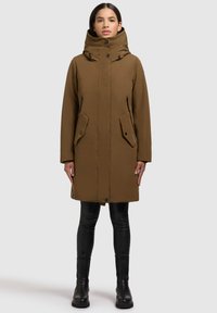 Parka marron avec un col haut, des poches zippées et des fermetures à pression. Fabriquée en tissu lisse, elle présente des manches longues et une coupe droite.