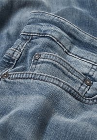 Denimtyg i en blekblå färg som har en framficka med svarta knappar och kontrasterande sömmar längs kanterna.