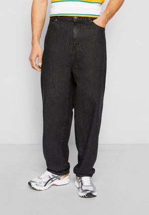 Mand iført løstsiddende sorte jeans, hvide sneakers og en stribet skjorte, med den ene hånd i lommen, stående på et gråt gulv.