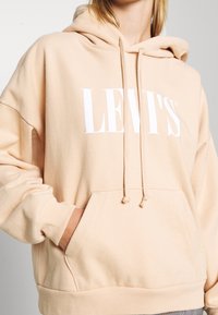 Felpa beige con logo bianco "LEVI'S", dotata di tasca frontale, cordini e un tessuto morbido e strutturato. Vestibilità rilassata con maniche lunghe.