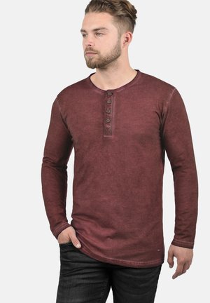 Langärmliges burgunderfarbenes Henley-Shirt mit fünf Knöpfen am Hals und einem strukturierten, leicht ausgebleichten Stoff. Kombiniert mit dunklen Jeans.