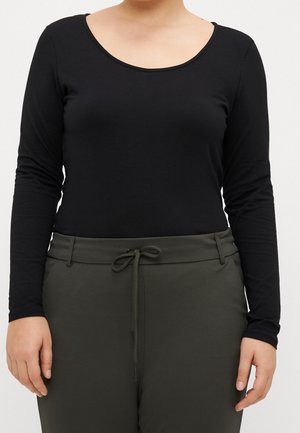 Person iført en sort, langærmet top med rund hals, stoppet ind i mørkegrønne bukser med bånd i taljen og bæltestropper.