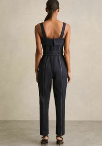Zwart denim jumpsuit met brede banden, getailleerde pasvorm, riemdetails en contrasterende witte stiksels. Glad textuur, rechte pijpen met enkel lengte.