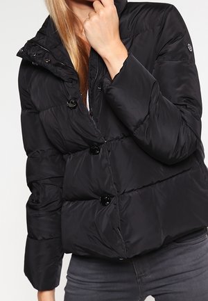 Kobieta w czarnej puchowej kurtce z dużymi guzikami, w ciemnoszarych dżinsach, stoi na tle jasnego, jednolitego tła.