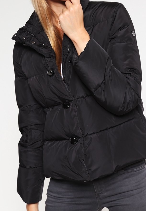 Femme portant une doudoune noire avec de gros boutons, associée à un jean gris foncé, debout devant un fond clair uni.