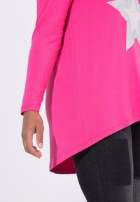 Rosa Langarm-Tunika mit asymmetrischem Saum, die ein silbernes Sternen-Design aufweist. Der Stoff wirkt weich und glatt und ergänzt schwarze Leggings.
