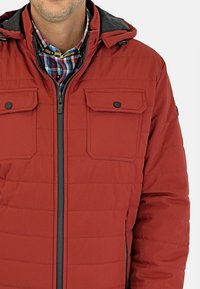 Chaqueta roja acolchada con cierre de cremallera, dos bolsillos en el pecho con solapas y una capucha negra. Presenta un tejido suave y texturizado.