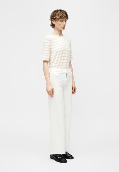 Άτομο με κοντά μαλλιά που φοράει μπεζ και άσπρη ριγέ κοντομάνικη μπλούζα, λευκό παντελόνι με φαρδιά πόδια και μαύρα μοκασίνια, στέκεται πλάγια.