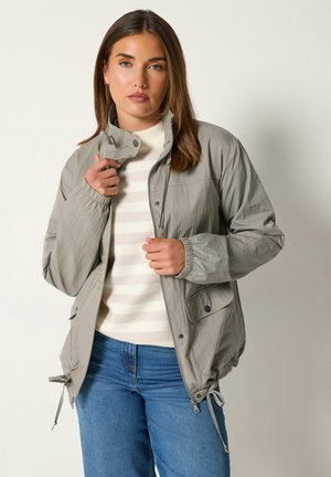 Femme ajustant le col d’une veste gris clair sur un pull rayé blanc et beige et un jean bleu, debout devant un fond clair uni.