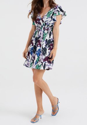 Nainen, joka pitää päällään lyhyttä kukkakuosista mekkoa, jossa on röyhelöiset hihat, ja siniset, remmillä varustetut korkokengät, seisten jalat ristissä.