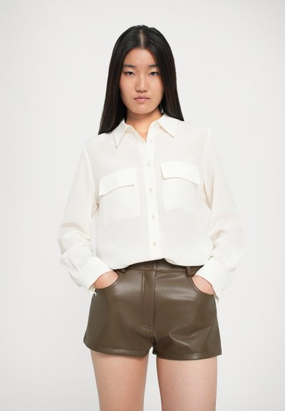 Fehér, hosszú ujjú ing, gombos záródással és két mellzsebbel, barna bőrnadrággal párosítva, amely fényes felülettel rendelkezik és oldalzsebekkel van ellátva.