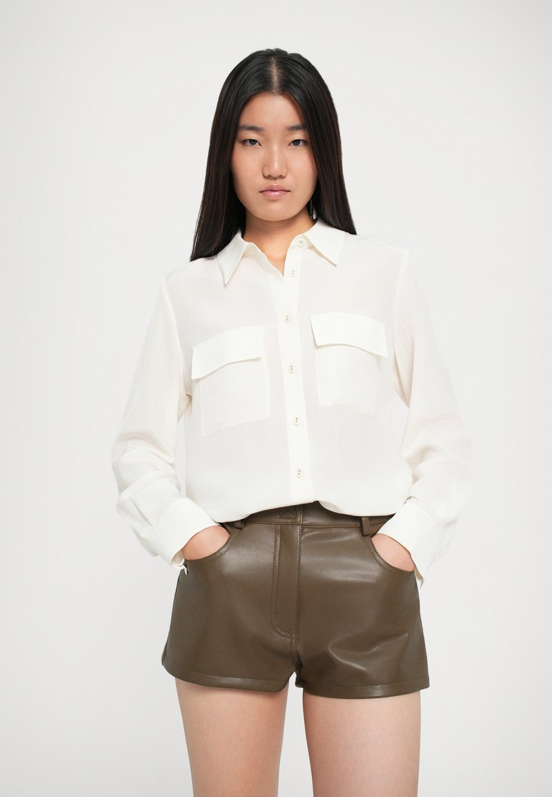 Camisa blanca de manga larga con cierre de botones y dos bolsillos en el pecho, combinada con pantalones cortos de cuero marrón con acabado brillante y bolsillos laterales.
