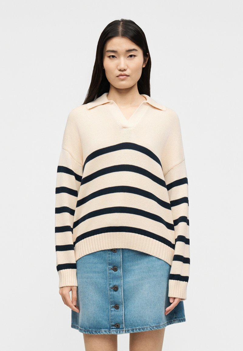 Γυναίκα που φοράει ένα εκρού πουλόβερ με οριζόντιες μπλε ρίγες και μια μπλε τζιν φούστα με κουμπιά, στέκεται μπροστά σε λευκό φόντο.