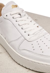 Bílé kožené tenisky s plochými šňůrkami, perforovanou špičkou, texturovanou gumovou podrážkou a zlatým logem na jazyku. Hladký povrch a minimalistický design.