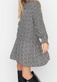 Schwarz-weiß gemustertes Kleid mit geometrischen Designs, langen Ärmeln, gerafftem Taillenbund und ausgestelltem Rock. Tragen mit kniehohen schwarzen Stiefeln.