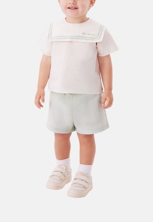 Un tout-petit portant une chemise rose pâle à manches courtes avec un col marin, un short vert clair, des chaussettes blanches et des baskets beiges à scratch, debout sur un fond blanc.