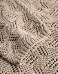 Könnyű bézs színű kötött takaró, amely texturált mintázattal rendelkezik, gyémánt alakzatokkal és nyitott varrás részletekkel, így lágy, kényelmes külsőt kölcsönöz.