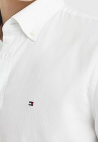 Wit overhemd met knopen en een spitse kraag; heeft een geborduurd logo in rood, blauw en wit op de linkerborst. Katoenen stoftextuur.