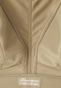 Sujetador deportivo beige con cierre de cremallera, diseño soportante y banda elástica. Incluye etiqueta de la marca "Champion Shock Absorber."