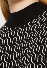 Svetr z pleteniny v černobílém vzoru s vysokým, žebrovaným límcem. Geometrický design obsahuje ostré úhly a jemnou textura.