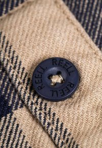 Botón negro de plástico redondo con "REELL" en relieve, sujeto a una tela beige con una trama texturada y un patrón de rayas azul oscuro.