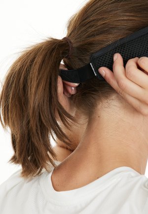 Person med brun hestehale, der fastgør et sort justerbart pandebånd bag sit hoved, iført en hvid skjorte.